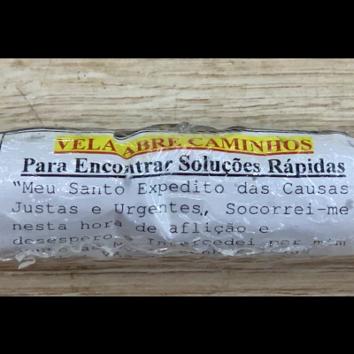 Vela artesanal amarela com pedaços verdes e vermelhos, embrulhada em plástico com etiqueta de oração, em madeira clara.