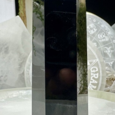 Obelisco de cristal preto polido hexagonal sobre superfície branca granular com fundo de cristais e símbolos astrológicos