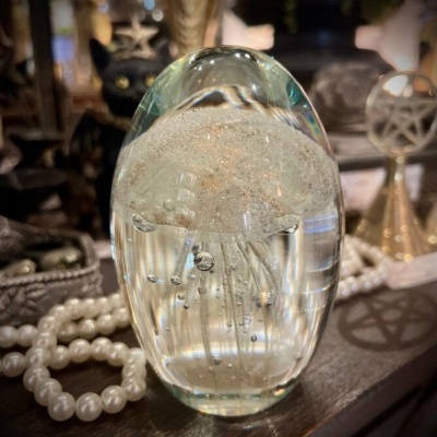 Objeto decorativo oval em vidro transparente com bolhas internas sobre mesa com colar de pérolas e objetos decorativos