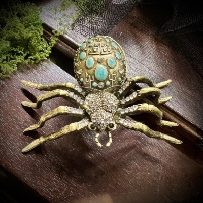 Broche de aranha dourado com pedras azuis e brilhantes sobre madeira escura e musgo verde