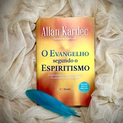 Livro O evangelho segundo o espiritismo de Allan Kardec sobre tecido branco com pena azul ao lado