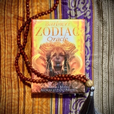 Baralho de cartas oracle Zodiac com rosário de contas de madeira e borla, sobre tecido listrado colorido