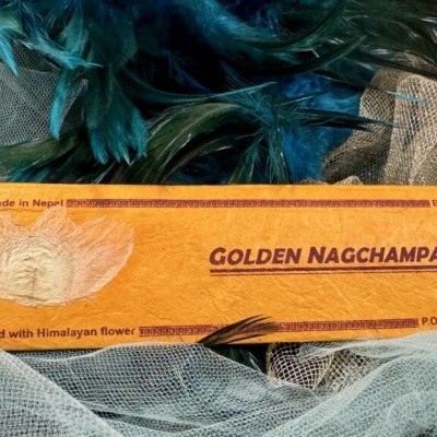 Embalagem amarela de incenso GOLDEN NAGCHAMPA com penas azuis e tecidos brancos ao redor