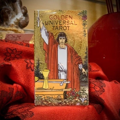 Caixa de tarot GOLDEN UNIVERSAL TAROT com figura de pessoa vestindo vermelho sobre tecido vermelho.