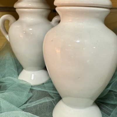 Dois potes brancos de porcelana com tampas sobre tecido verde translúcido.