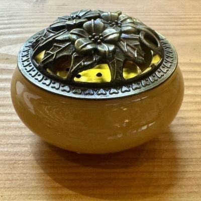 Vaso redondo amarelo com tampa metálica decorativa floral sobre mesa de madeira