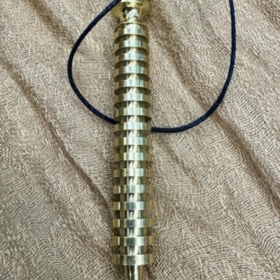 Pêndulo metálico dourado cilíndrico com cordão preto sobre tecido bege texturizado