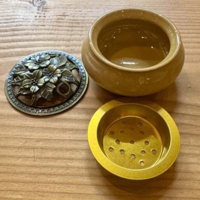Recipiente amarelo dourado, cerâmica castanha e peça metálica decorativa com flores em relevo sobre madeira