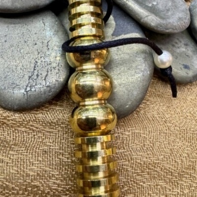 Pêndulo de metal dourado com cordão preto e conta branca sobre pedras e tecido