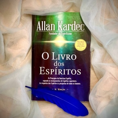 Livro O Livro dos Espíritos junto a tecido branco e pena azul