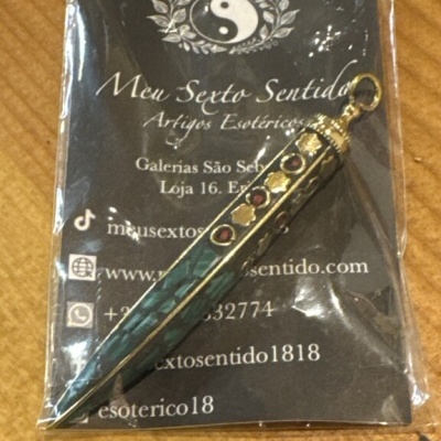 Pingente dourado com pedras coloridas em embalagem preta com texto branco
