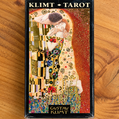 Caixa preta de cartas de tarot com ilustração colorida de Gustav Klimt e texto na embalagem em cima de madeira.