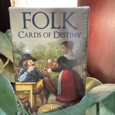 Pacote de cartas FOLK CARDS OF DESTINY com ilustração antiga entre folhas verdes.
