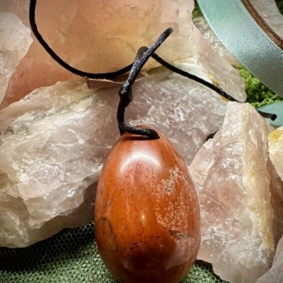 Colar com pendente oval de pedra vermelha e cordão preto sobre pedras de quartzo rosa e tecido verde.