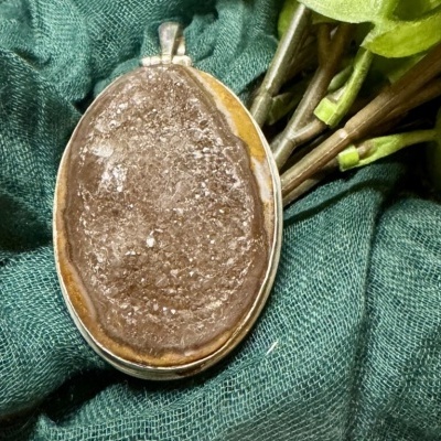 Pingente oval de pedra natural castanha com moldura prateada sobre tecido verde