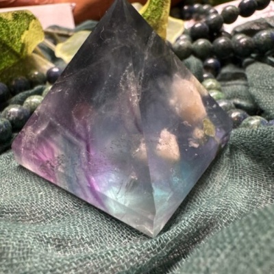 Pirâmide de cristal multicolor sobre tecido verde com contas e folhas ao fundo