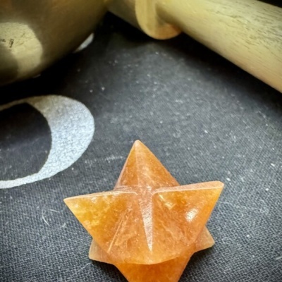 Cristal laranja em forma Merkaba sobre tecido preto com padrão branco ao lado de objeto metálico e bastão de madeira