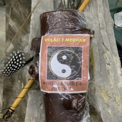 Vela castanha com etiqueta yin-yang embalada em plástico sobre pedras, com pena e varinha decorativa ao lado.