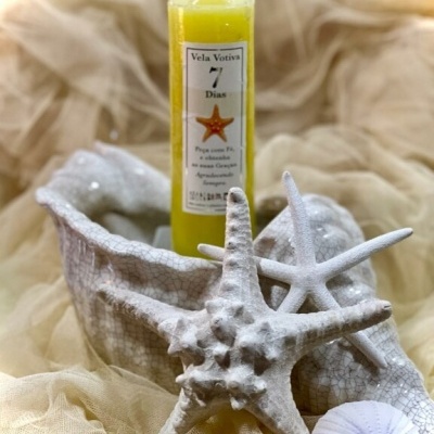 Vela votiva amarela com rótulo branco dentro de um objeto decorativo branco com estrelas-do-mar brancas ao redor, sobre tecido bege.
