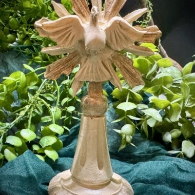 Escultura dourada de pomba com asas abertas sobre base redonda, decorada com folhagem verde ao fundo