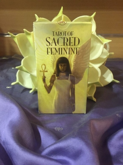 Caixa de tarô TAROT OF SACRED FEMININE com imagem de mulher e asas sobre tecido roxo e flores brancas.