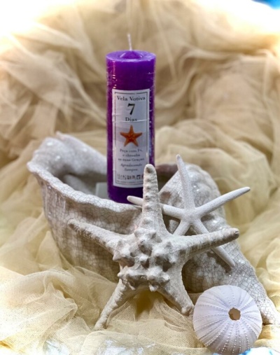 Vela votiva roxa em concha decorativa branca com estrelas-do-mar e ouriço-do-mar em tecido bege.