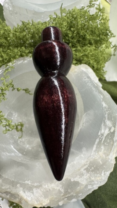 Figura feminina esculpida polida castanha avermelhada sobre cristais brancos com vegetação verde.