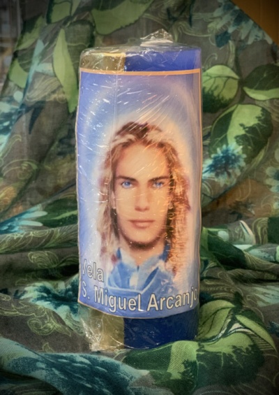 Vela azul e amarela com rótulo de São Miguel Arcanjo sobre tecido verde com padrão de folhas