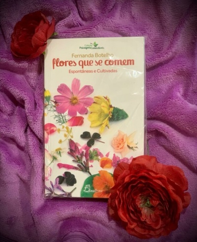 Livro com capa branca e flores coloridas sobre tecido roxo com flores vermelhas artificiais.