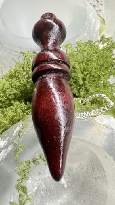 Pingente em madeira castanho escuro com corrente prateada sobre pedras e folhas verdes