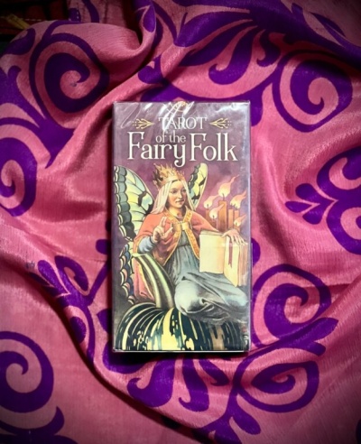 Caixa de tarot com uma fada ilustrada, sobre tecido rosa e roxo.