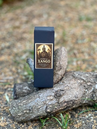 Caixa preta com rótulo dourado de produto chamado FLUIDO DE XANGÔ sobre troncos de madeira ao ar livre