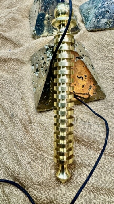 Pêndulo dourado espiral metálico com cabo preto sobre tecido bege com pirâmides de vidro