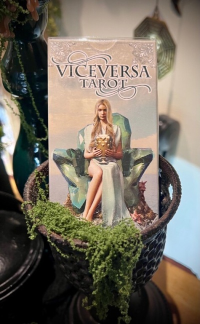 Caixa de tarô VICEVERSA TAROT com imagem de mulher loira sentada em cadeira verde, dentro de cesta com musgo