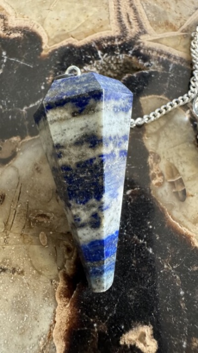 Pendente de pedra azul e branca com corrente prateada sobre fundo de pedra marrom e preto