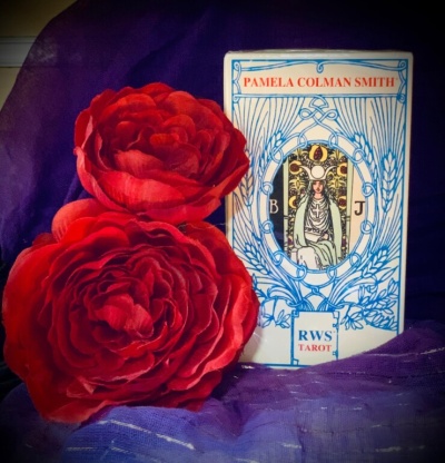 Caixa de cartas de tarot com texto e duas flores vermelhas artificiais junto a tecido roxo.