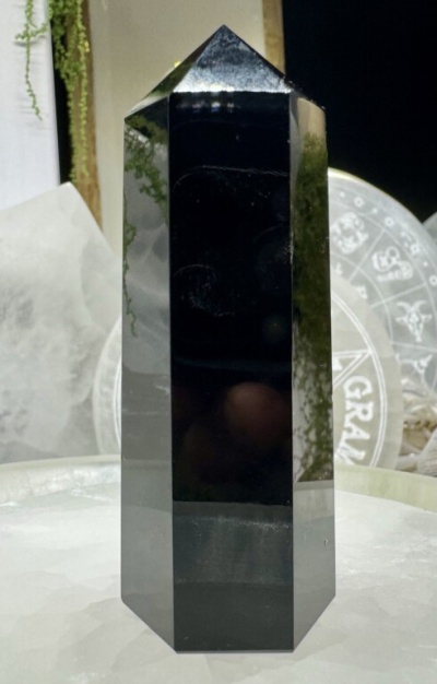 Obelisco de cristal preto polido hexagonal sobre superfície branca granular com fundo de cristais e símbolos astrológicos