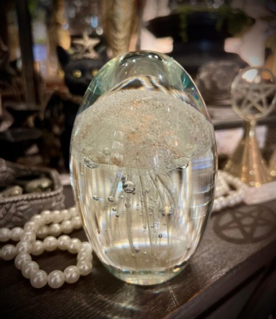Objeto decorativo oval em vidro transparente com bolhas internas sobre mesa com colar de pérolas e objetos decorativos