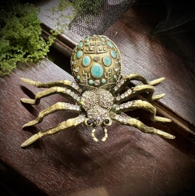 Broche de aranha dourado com pedras azuis e brilhantes sobre madeira escura e musgo verde