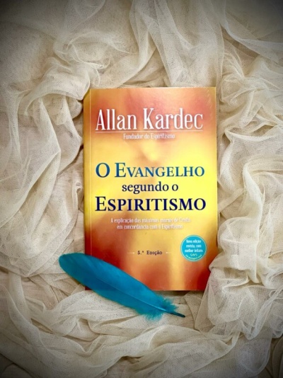 Livro O evangelho segundo o espiritismo de Allan Kardec sobre tecido branco com pena azul ao lado