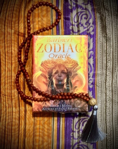 Baralho de cartas oracle Zodiac com rosário de contas de madeira e borla, sobre tecido listrado colorido