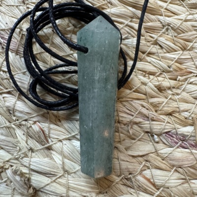 Colar com pendente de pedra verde e cordão preto sobre tecido entrelaçado.