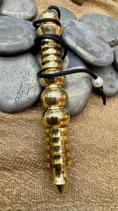 Pêndulo de metal dourado com cordão preto e conta branca sobre pedras e tecido