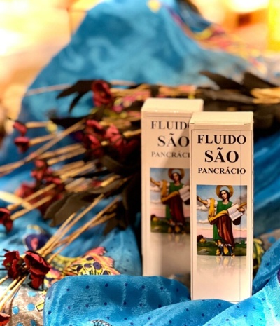 Duas caixas brancas de fluido São Pancrácio com imagem de santo, sobre tecido azul e flores secas vermelhas e pretas.