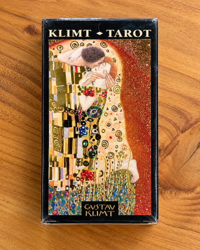 Caixa preta de cartas de tarot com ilustração colorida de Gustav Klimt e texto na embalagem em cima de madeira.