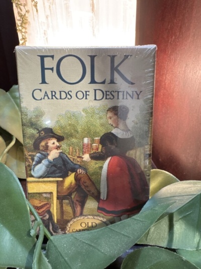 Pacote de cartas FOLK CARDS OF DESTINY com ilustração antiga entre folhas verdes.