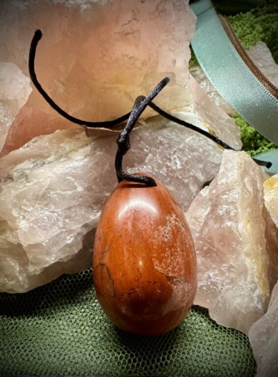 Colar com pendente oval de pedra vermelha e cordão preto sobre pedras de quartzo rosa e tecido verde.