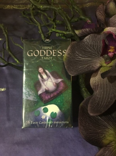 Caixa de tarot Triple Goddess ao lado de flores roxas e galhos verdes sobre tecido roxo.