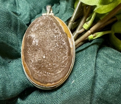 Pingente oval de pedra natural castanha com moldura prateada sobre tecido verde