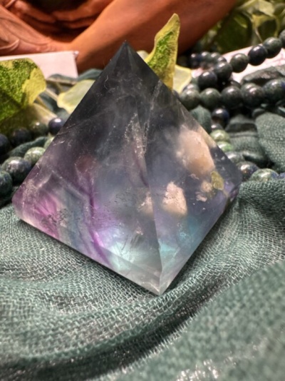 Pirâmide de cristal multicolor sobre tecido verde com contas e folhas ao fundo